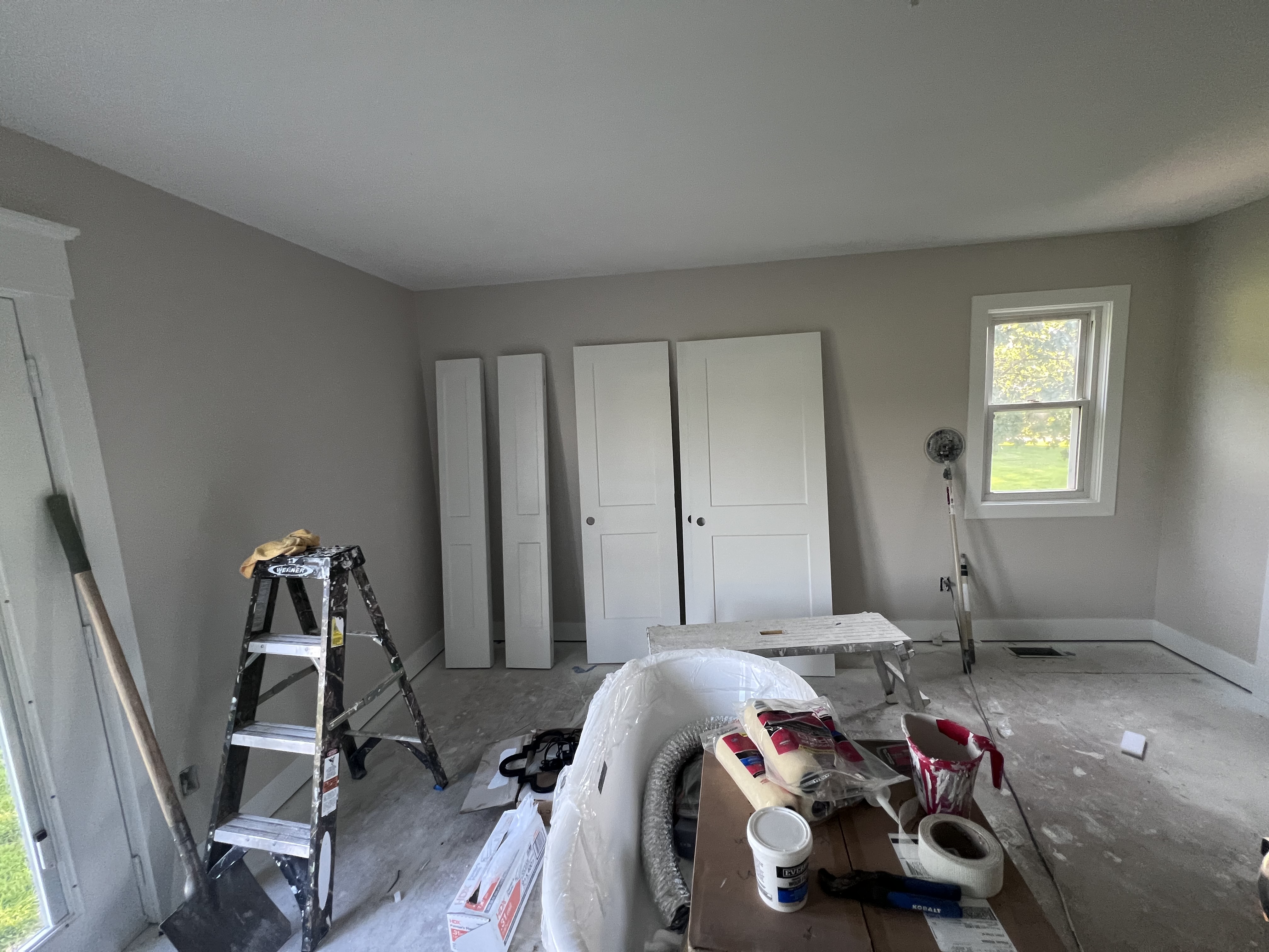 A gray-painted bedroom with white closet doors leaning against the wall and a ladder in the middle of the room
