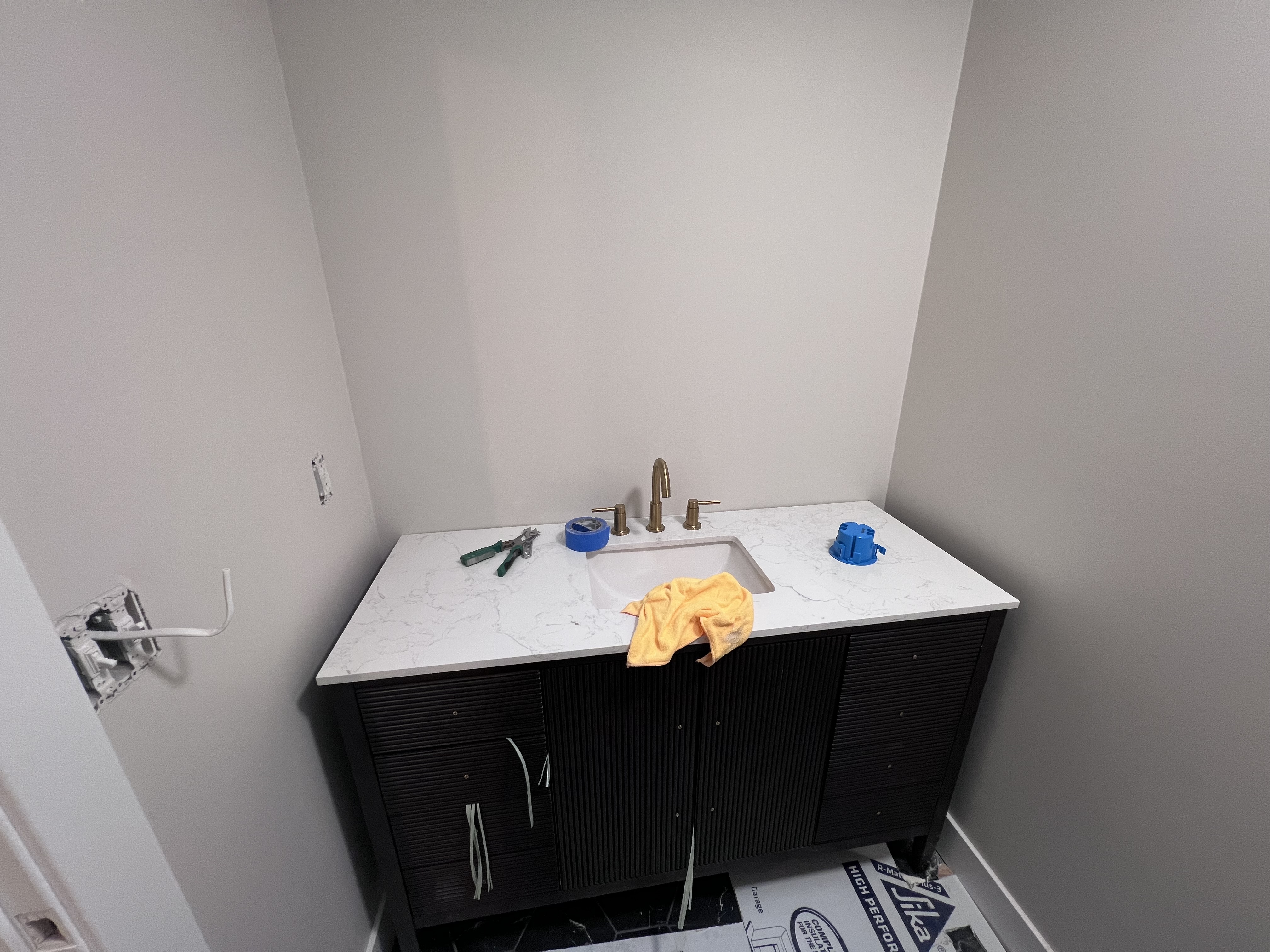 A guest bathroom vanity with a black wood cabinet, white quartz countertop, brass faucet, and paint supplies still on the counter