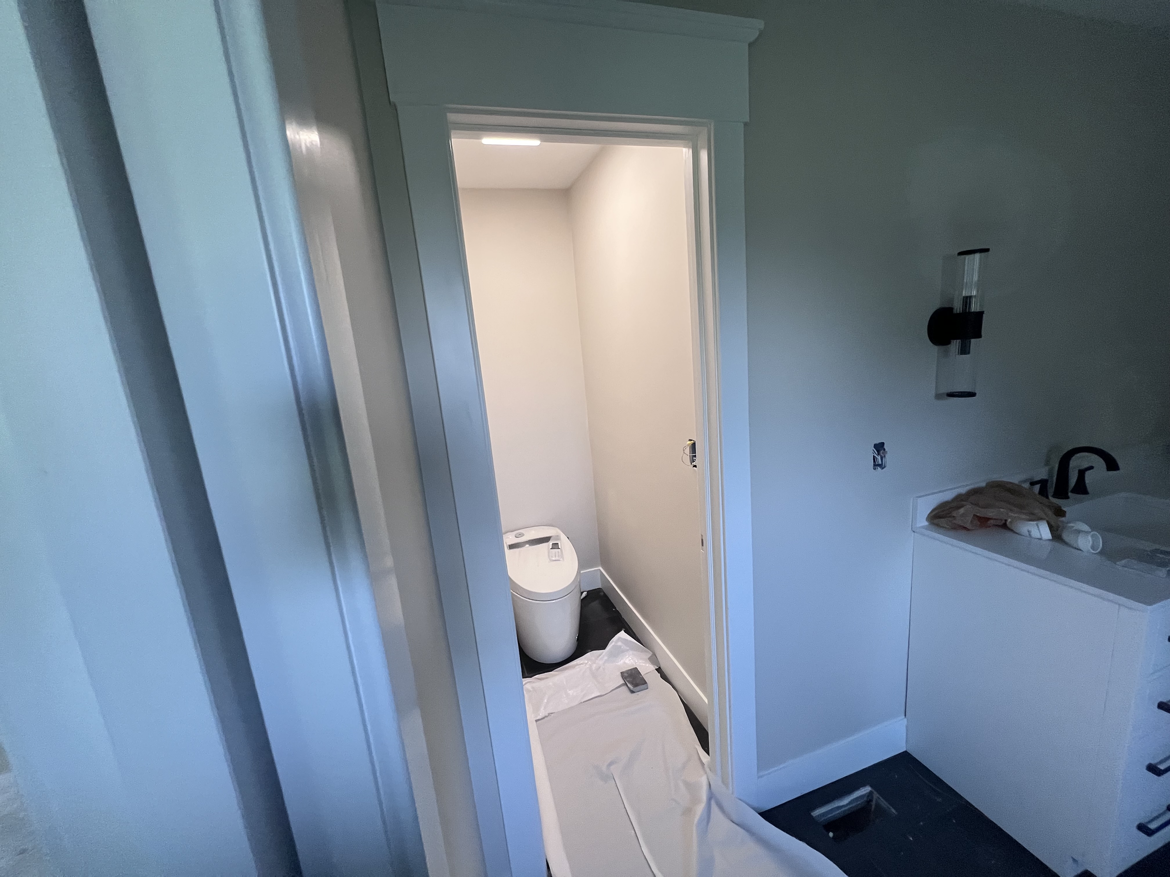 A view of the main suite bathroom from the vanity side, with a white vanity in the foreground, modern black sconce on the wall, and a toilet visible through the doorway