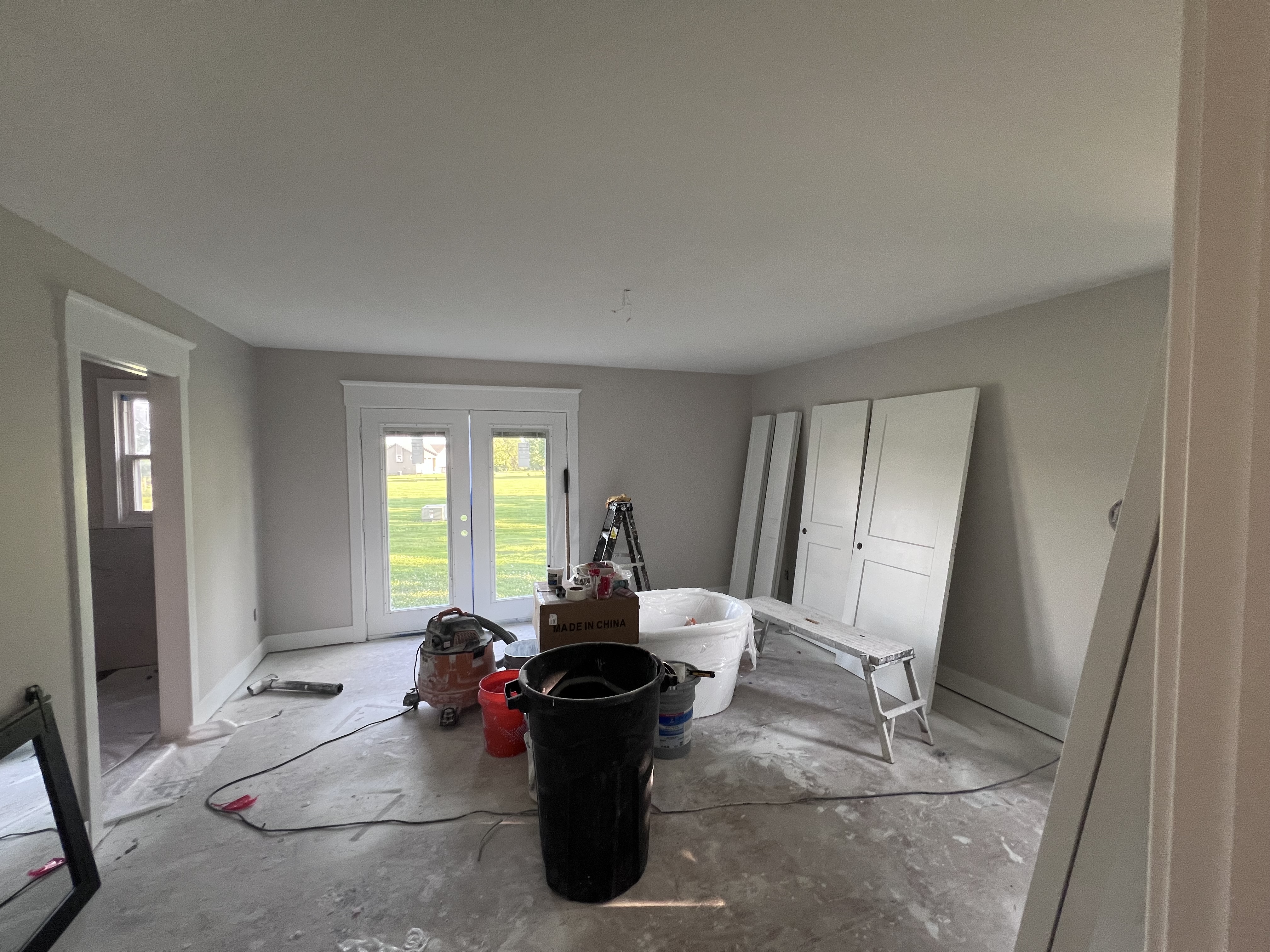 Master bedroom with freshly painted gray walls, French doors looking out at the yard, paint cans and a ladder in the middle of the room