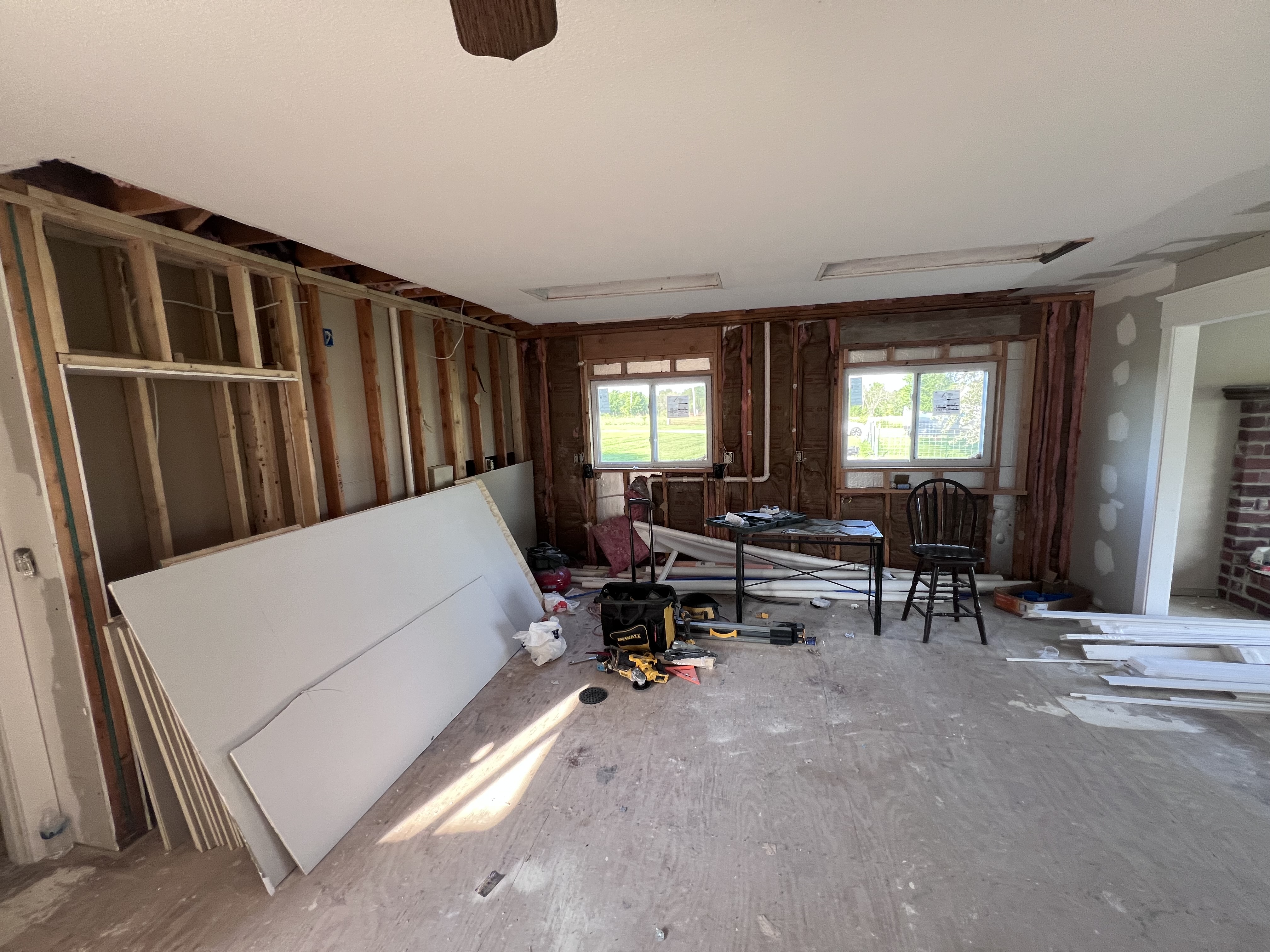 A gutted room with wall studs exposed, drywall sheets on the floor, two windows looking out at the yard, and tools scattered around