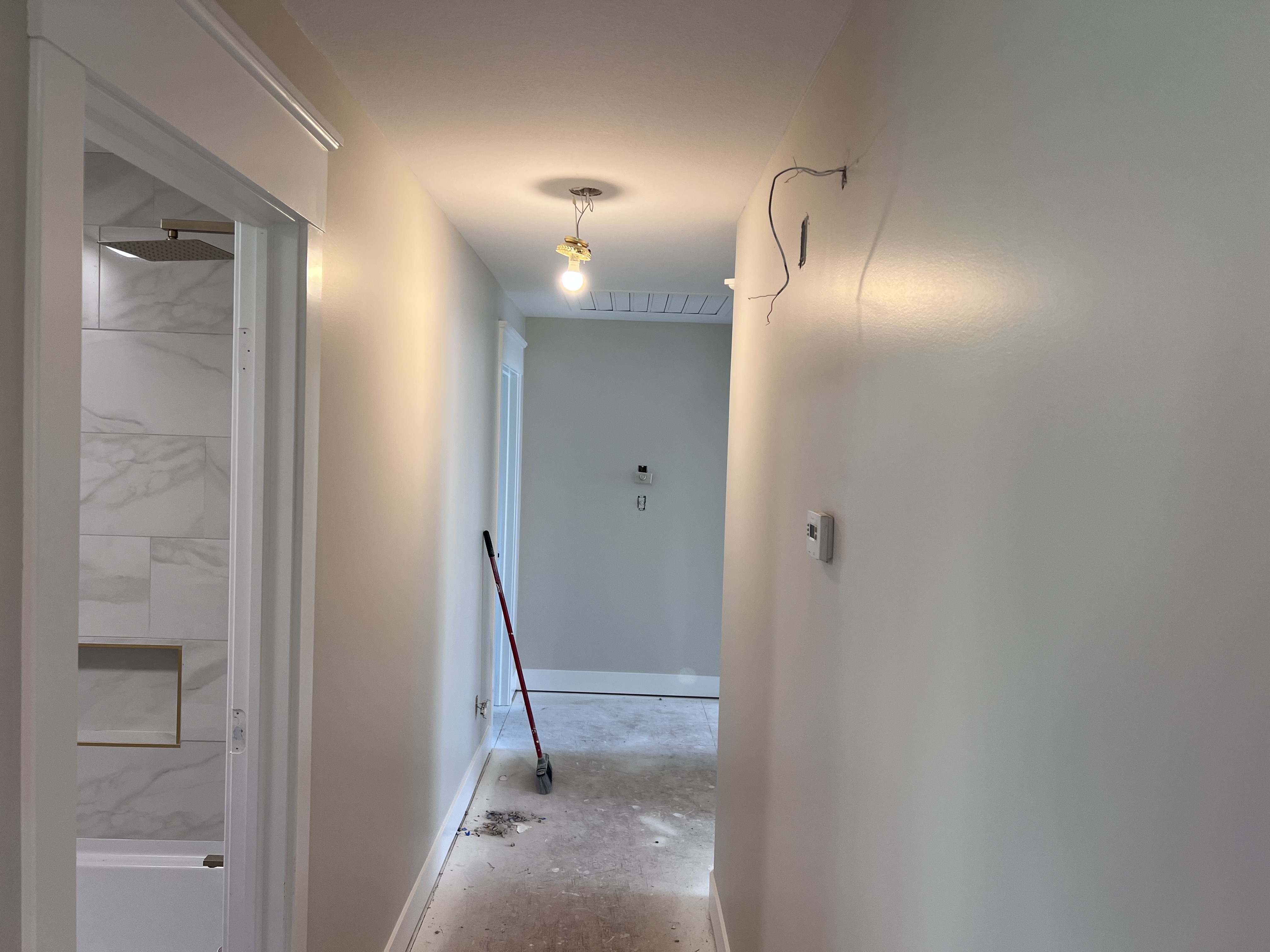 A freshly painted hallway in light blue-gray, the bathroom door open on the left, a single light bulb hanging from the ceiling, a broom on the floor