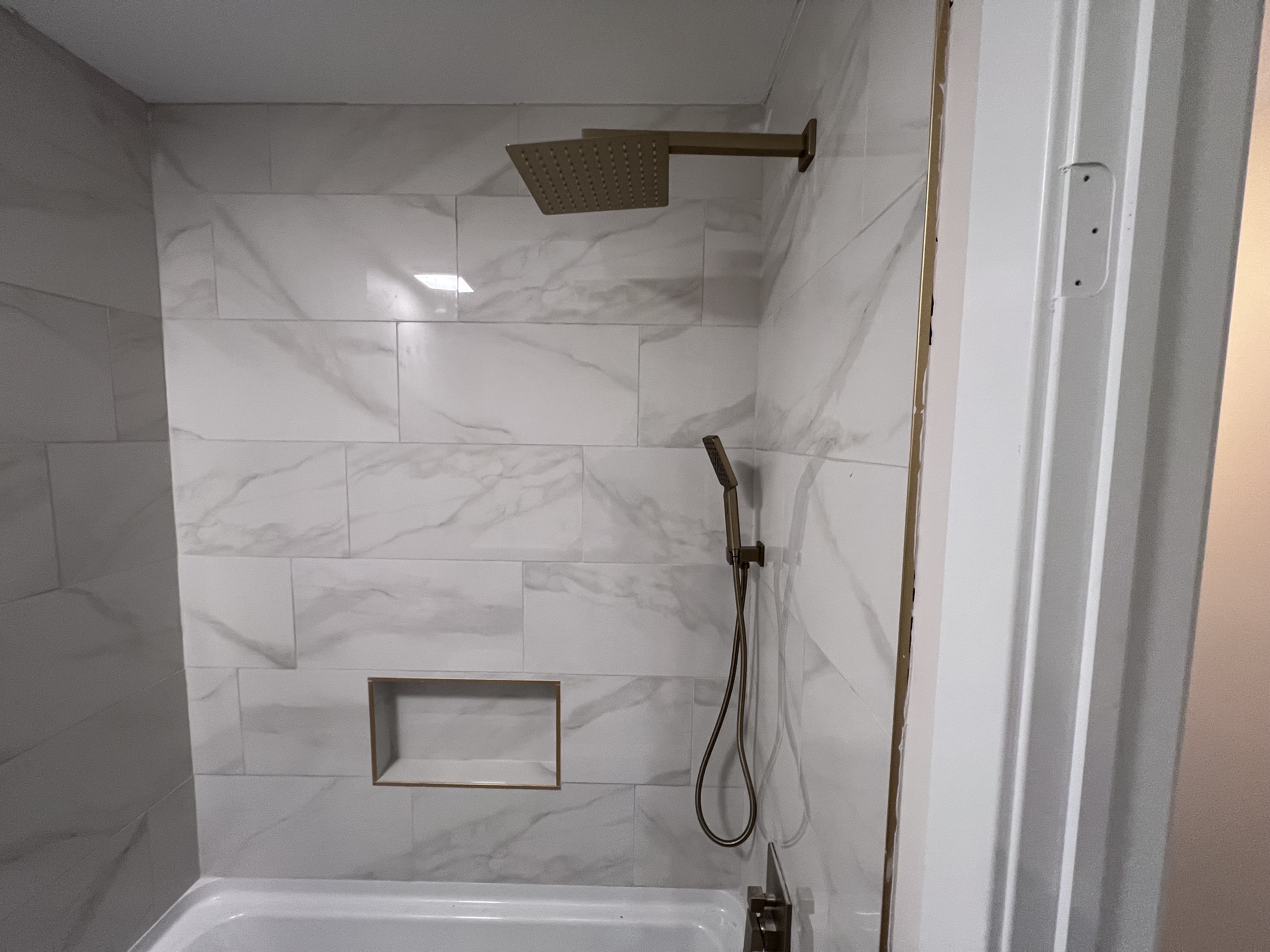 A shower with marble-pattern tile, gold square rainhead, gold handheld sprayer, and a built-in tile niche